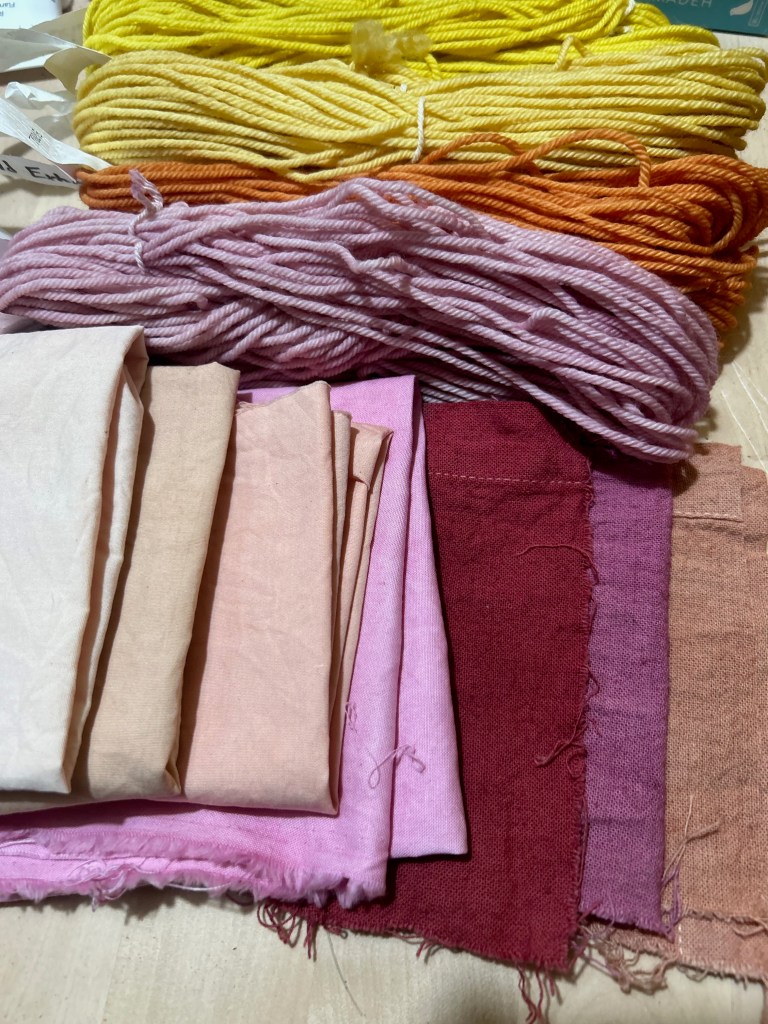 A display of folded fabrick squares and small skeins of yarn dyed pinks, yellows, and oranges. 