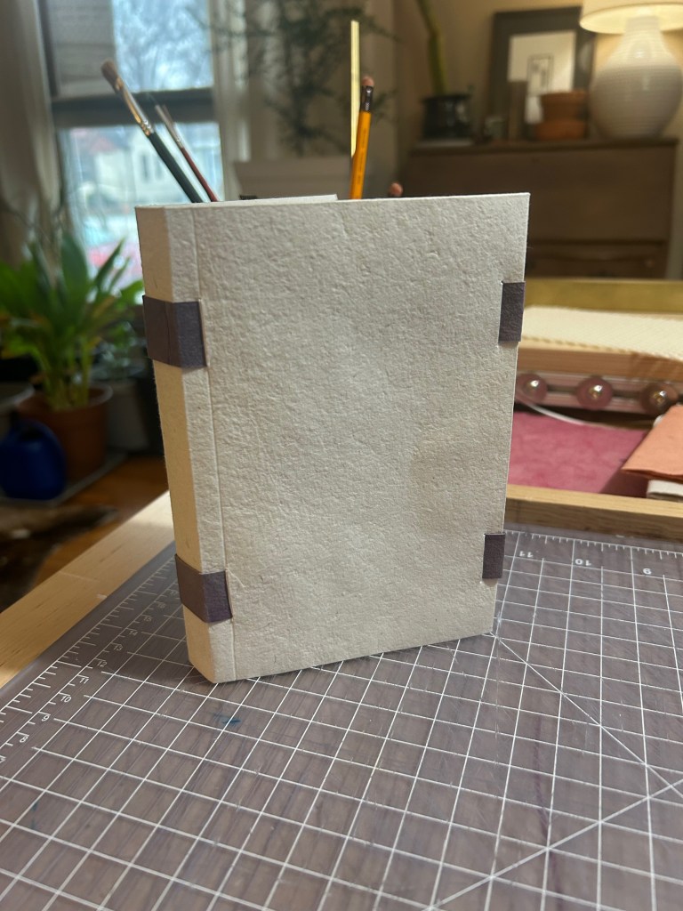 A white book with visible sewing tapes at the spine and fore edge is standing upright on a cutting mat.