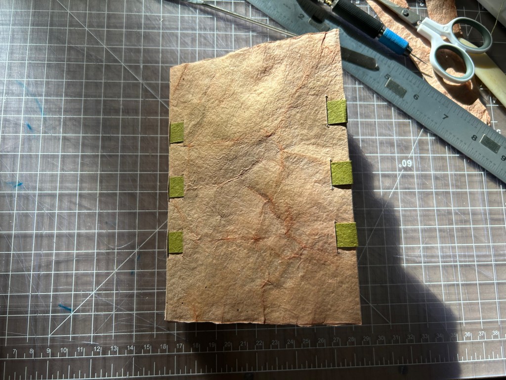 A moody picture of a tan book with visible green support tapes at the spine and fore edge is laying on a cutting mat.